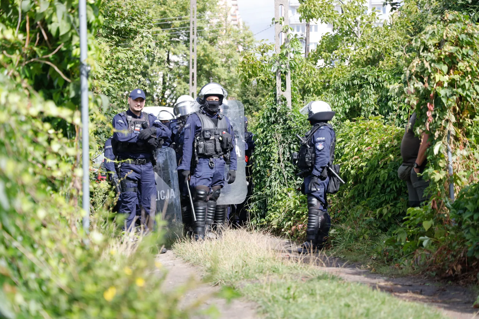 Funkcjonariusze oddziałów prewencji w kaskach z tarczami wchodzą wąską alejką