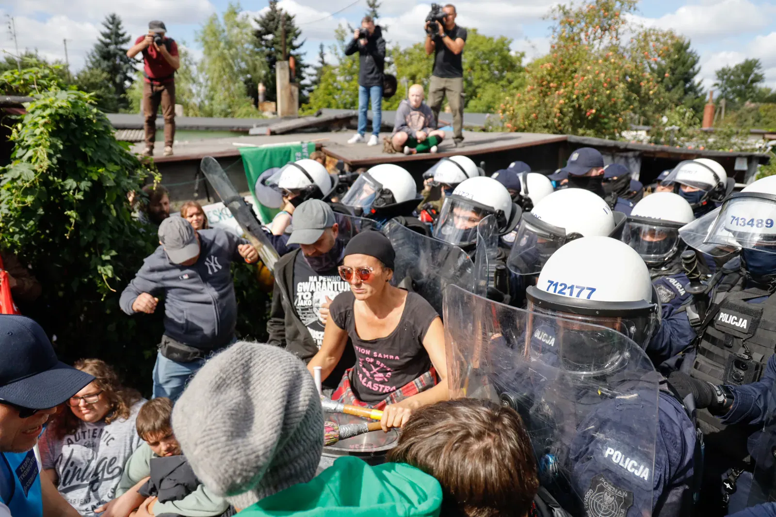 Natarcie policyjnych oddziałów prewencji na protestujących mieszkańców i aktywistów spychanych tarczami; z dachów sąsiednich budynków media i uczestnicy nagrywają zdarzenie. Osiedle Maltańskie w Poznaniu