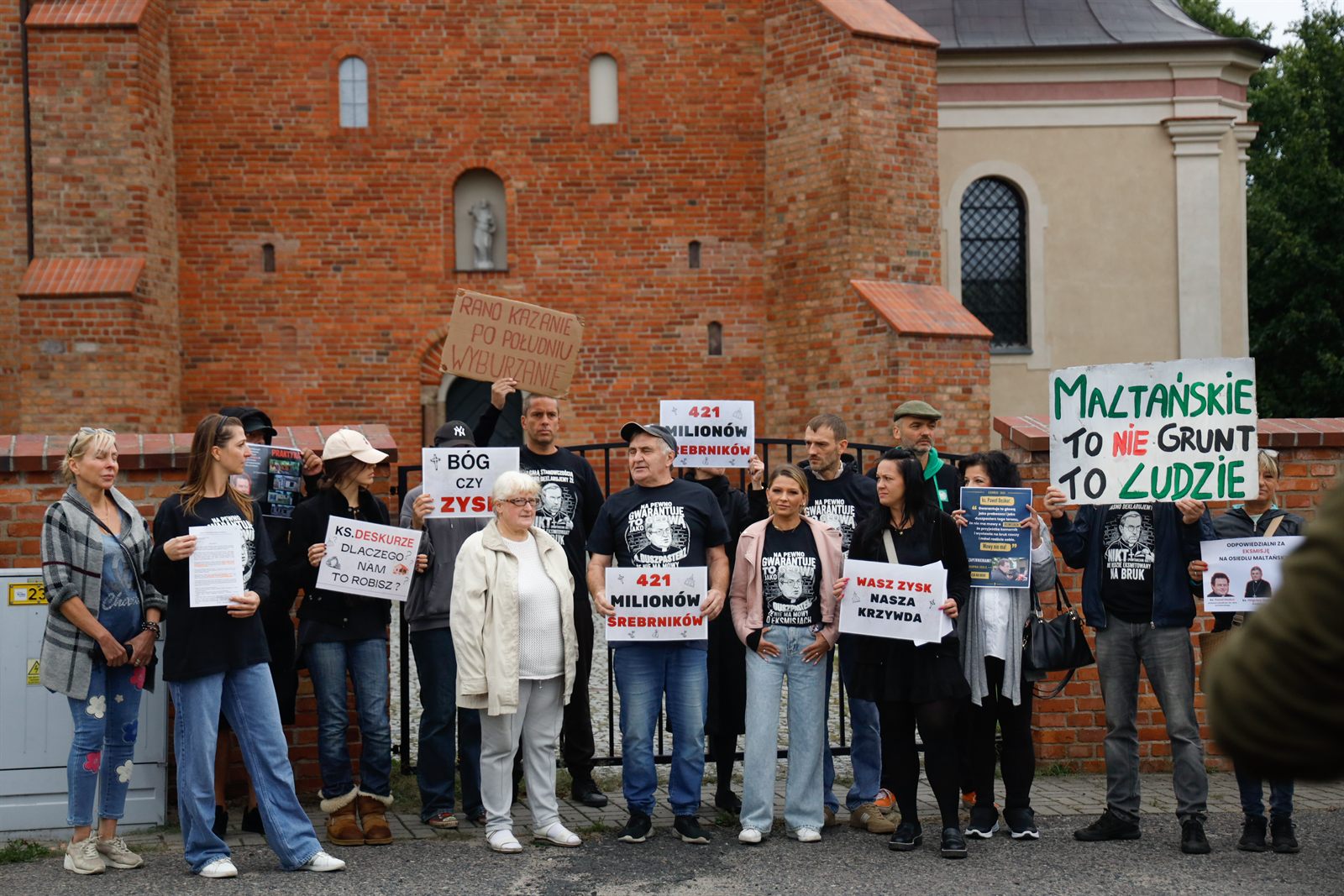 Kilkanaście osób z tablicami stoi przed ceglanym kościołem, na tablicach napisy „Bóg czy zysk?”, „421 milionów srebrników”, „Maltańskie to nie grunt, to ludzie”