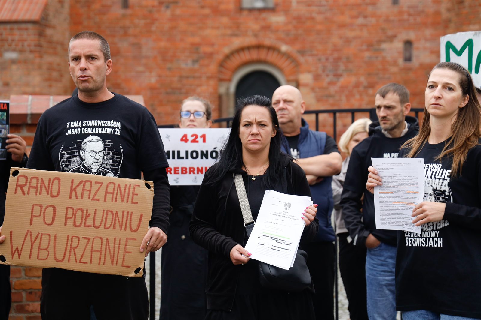 Mężczyzna trzyma tekturową tablicę z napisem „Rano kazanie, po południu wyburzanie”, obok kobieta z kartką „Wasz zysk — nasza krzywda”