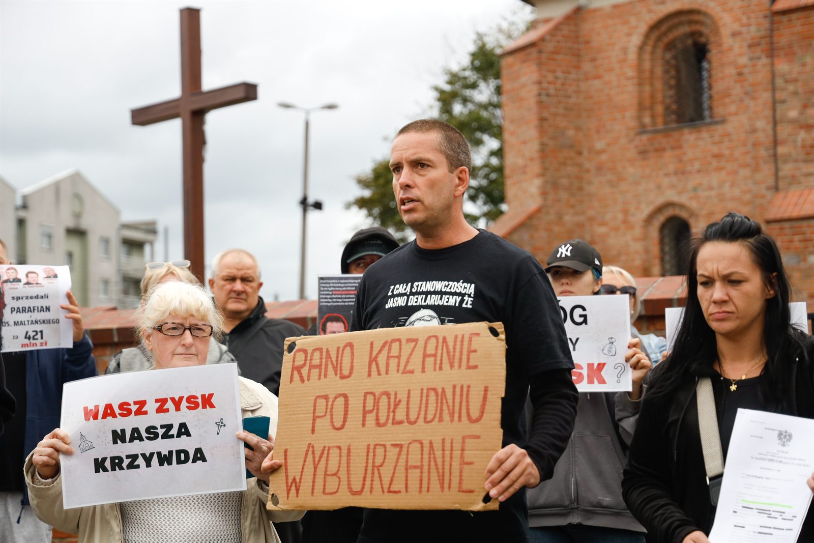 Mężczyzna z tekturową tablicą „Rano kazanie, po południu wyburzanie” i kobieta z kartką „Wasz zysk — nasza krzywda” stoją pod krzyżem przed kościołem św. Jana Jerozolimskiego za Murami w Poznaniu