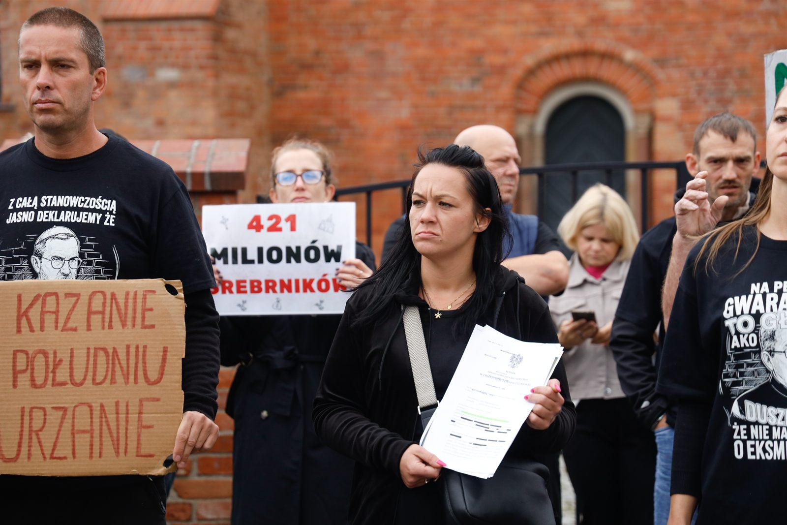 Kobieta trzyma w dłoniach kilkustronicowy dokument sądowy z godłem, w tle uczestnicy protestu