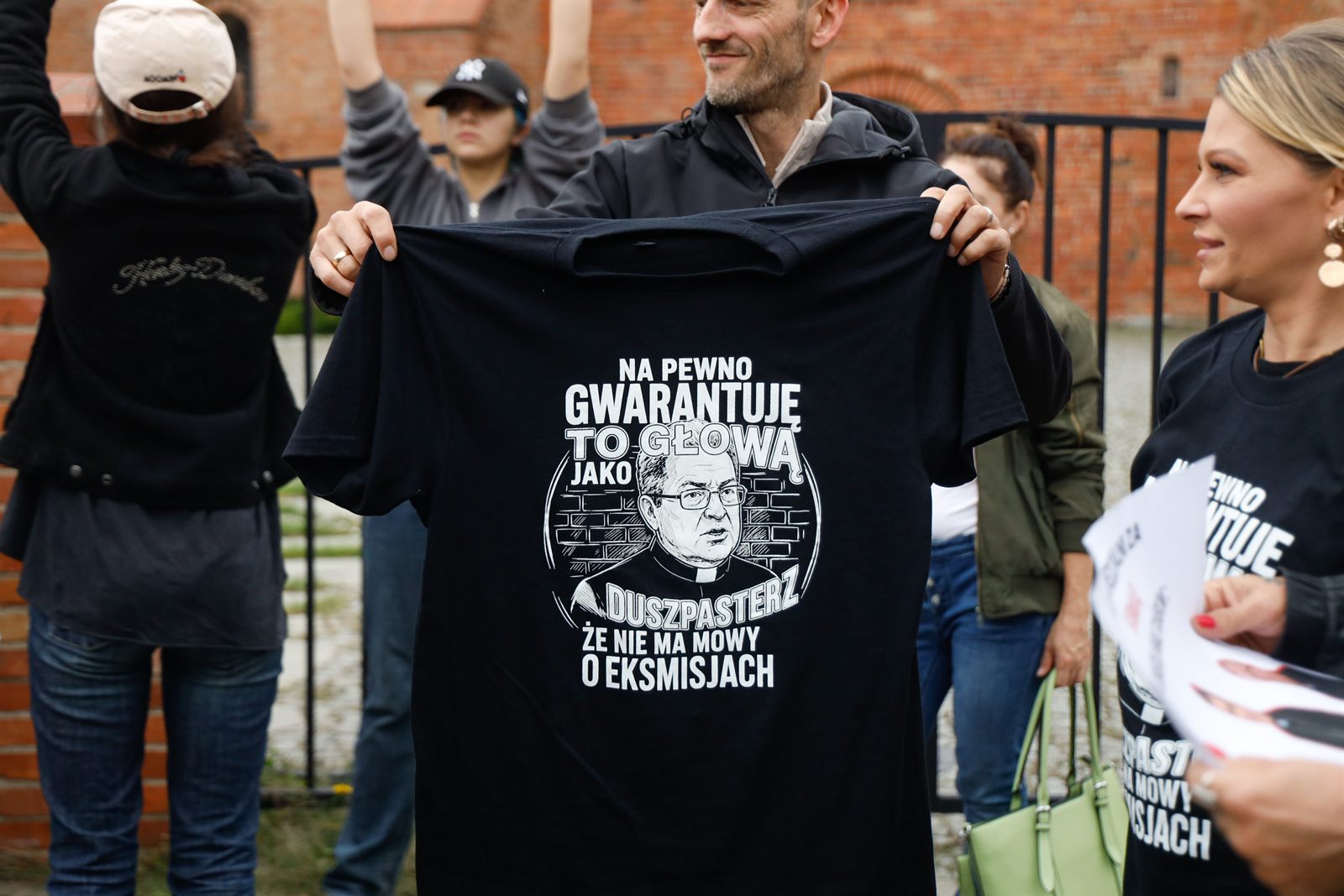 Mężczyzna rozkłada przed sobą czarną koszulkę z podobizną duchownego i napisem „Na pewno gwarantuję to głową jako duszpasterz, że nie ma mowy o eksmisjach”