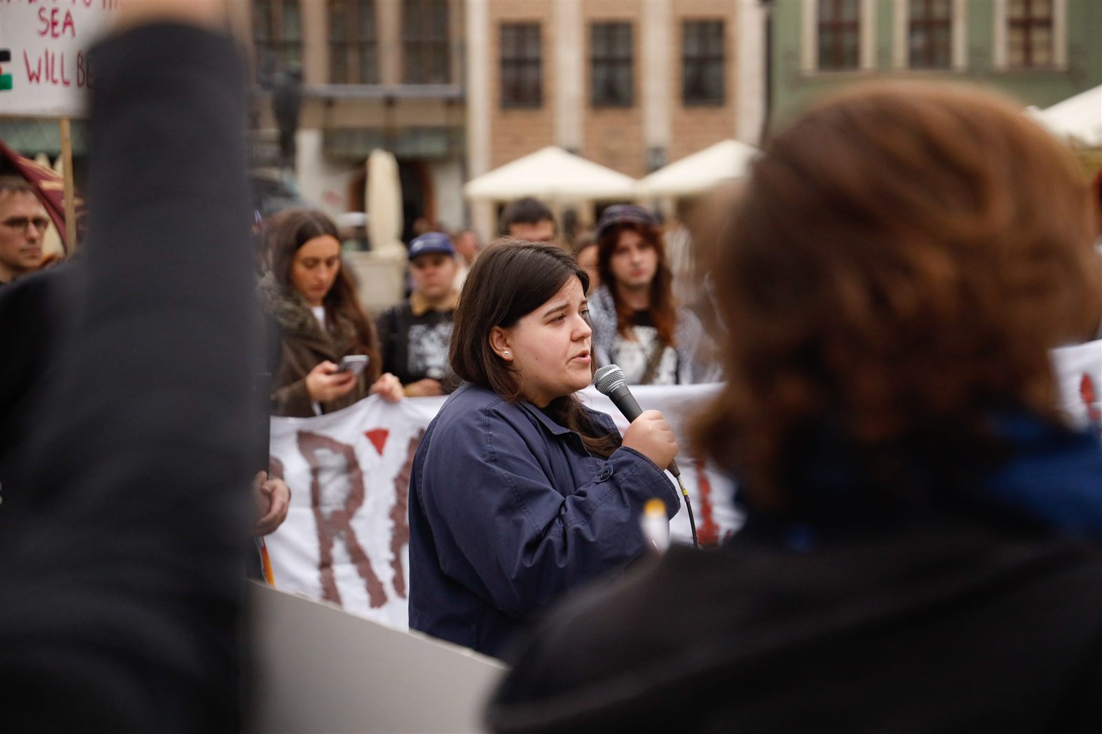 Anna Pieciul przemawia do mikrofonu, za nią uczestnicy i transparent