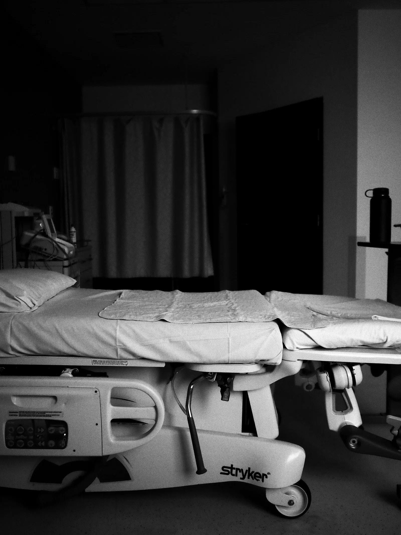 Czarno-białe zdjęcie pustego łóżka szpitalnego na kółkach w zaciemnionej sali, w tle zasłona i aparatura medyczna