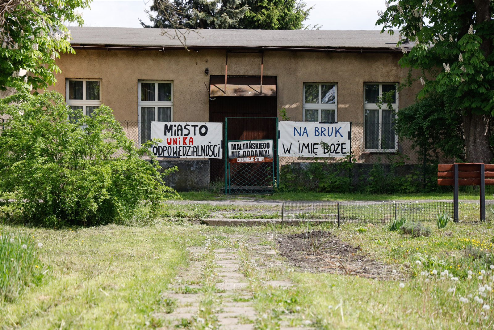 Parterowy budynek świetlicy na osiedlu Maltańskim z zamkniętą bramą; na ogrodzeniu transparenty "Miasto unika odpowiedzialności", "Maltańskiego nie oddamy! Eksmisje stop!" i "Na bruk w imię Boże"
