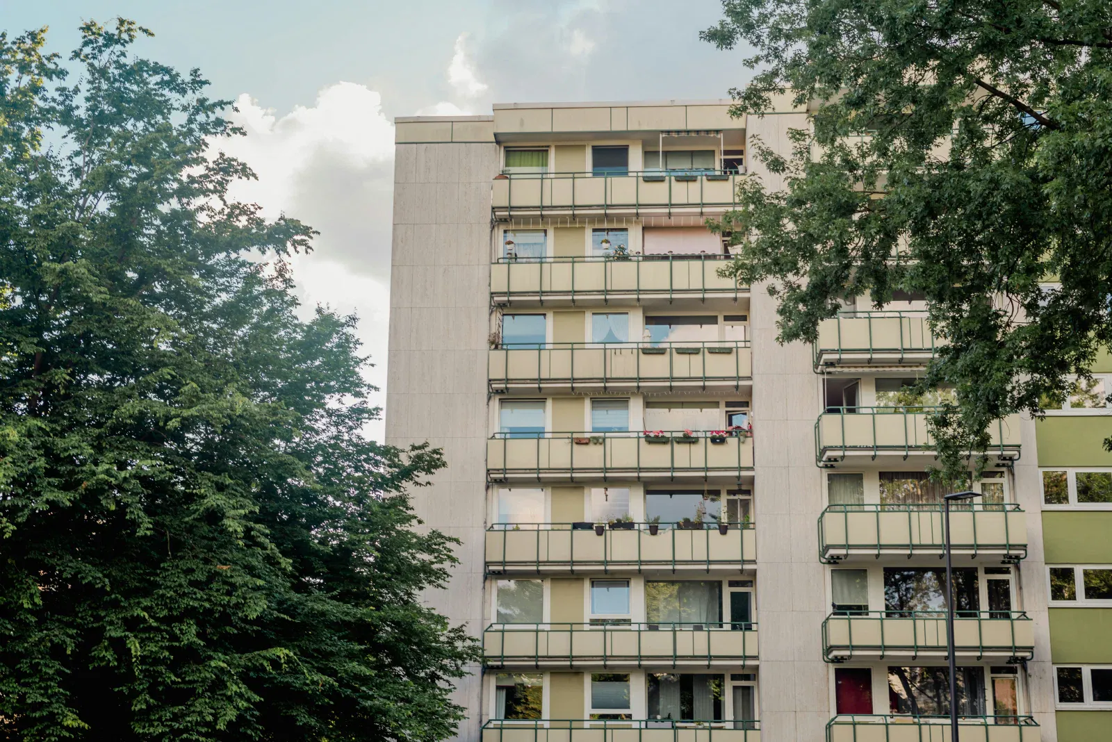 Szaro-beżowy budynek wielokondygnacyjny z balkonami, po lewej stronie drzewo
