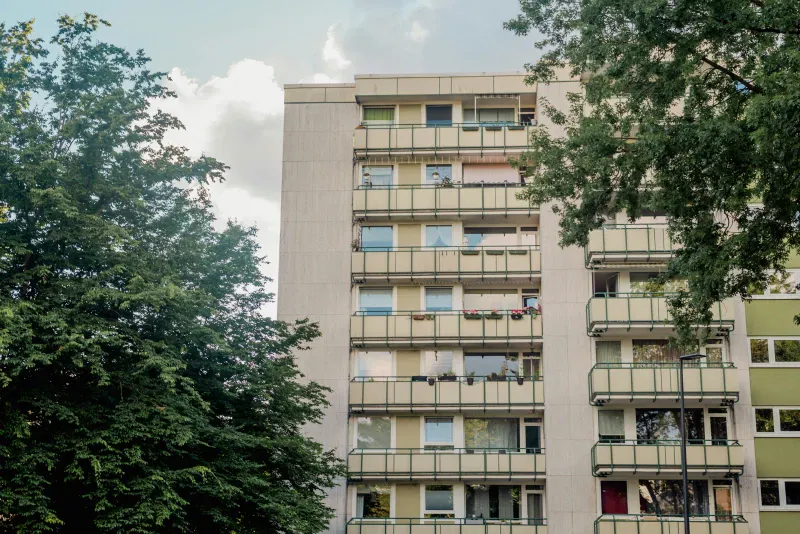 Szaro-beżowy budynek wielokondygnacyjny z balkonami, po lewej stronie drzewo