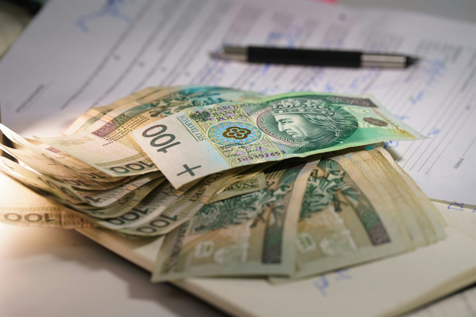 Plik banknotów stuzłotowych leży na wypełnionych formularzach obok długopisu